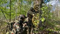 Zwei Soldaten befinden sich in einem Wald, hinter einem Baum und halten ihr G36 im Anschlag