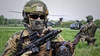 On a green field, two soldiers – one of them with a dog – kneel in front of two helicopters in the landing zone.