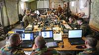 Numerous soldiers sit in a large tent at tables with computers.