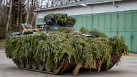 An IFV camouflaged with green branches moving on a road between hangars.