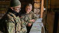 Two officers are standing in a hut in front of maps spread out on the window sill and negotiate with each other.