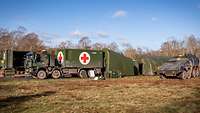 Several green tents, military vehicles and containers with the red cross symbol are standing in an open area.