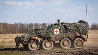 A camouflaged wheeled armoured medical vehicle with a red cross in a white circle is speeding across a field.