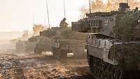 Column of several tanks moves across muddy ground.