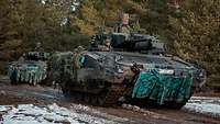 Two tanks move along the edge of a forest in single file. There is some snow in the foreground.