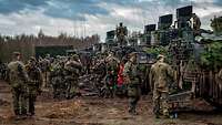 Numerous IFVs are lined up side by side in brown mud. Numerous soldiers stand behind the vehicles' rear doors.