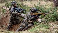 Two soldiers aiming their rifles kneel in a position located in a wood.