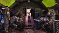 Inside a green tent, four medics treat two wounded soldiers on raised stretchers. 