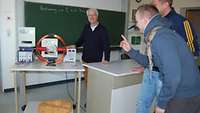 Drei Männer sind in einem Klassenzimmer und stehen vor einem Gerät, das Stromspannungen misst.