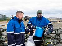 Zwei Soldaten stehen mit einem blauen Eimer an einer Pipeline mit einem Entnahmerohr. 