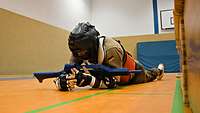 Ein Soldat bewegt sich robbend auf dem orangefarbenen Hallenboden mit einem blauen Übungsgewehr.