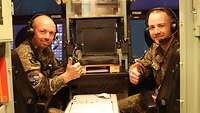 Zwei Soldaten sitzen zusammen in einer Kabine. Im Hintergrund befinden sich Bildschirme und technisches Gerät.
