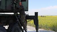 Ein Soldat steht auf einer Leiter und prüft etwas an dem Radar, im Hintergrund Felder und Wiesen.