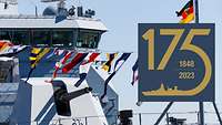 Blick auf das Vorschiff eines grauen Kriegsschiffs; bunte Signalflaggen wehen von einer langen Leine.