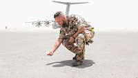 Ein Soldat hebt etwas vom Boden auf. Im Hintergrund ein Flugzeug, das auf einem Flugplatz steht.