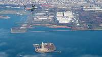 A plane flies over a city by the sea, a small island with a large statue in the apron.