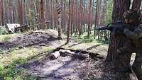 Ein Soldat läuft im Wald mit dem Gewehr im Anschlag auf eine Stellung mit einer Kanone zu.