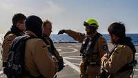 Der Flugdeckoffizier instruiert auf dem Flugdeck die Soldaten. Im Hintergrund das Flugdeck, dahinter das Meer. 