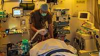 Ein Anästhesist setzt einem Patienten eine Beatmungsmake auf, im Hintergrund viele medizinische Geräte. 