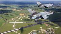 Two Tornado aircraft fly in formation in the sky. Below them, the Jagel airfield can be clearly seen.