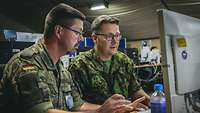 Two soldiers in green uniforms sit in front of a monitor.