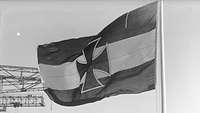 Schwarz-Weiß-Aufnahme einer dreifarbig-längsgestreiften Flagge mit einem Eisernen Kreuz in der Mitte