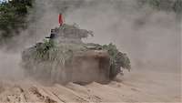 Ein Panzer mit roter Flagge fährt schnell einen sandigen Übungsplatz, Sand wirbelt auf.