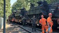 Soldaten und Zivilisten in orangefarbener Schutzkleidung verladen Panzer auf Zugwaggons. 