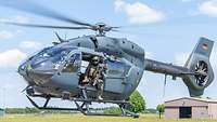 An H145M helicopter hovers above the ground. A soldier with a rifle sits at the door.