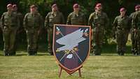 Hinter einem Kompaniewappen auf einem Ständer stehen Soldaten in Formation.