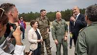 Politicians and military officers stand together on a tarmac and address the press.