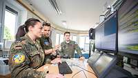 Three soldiers stand in front of monitors and discuss a mission.