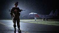 A soldier stands on the airfield with rifle at night and rain. Behind him is a fighter jet.