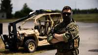 A masked soldier is standing in front of a support vehicle.