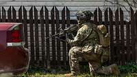 Ein US-Soldat hockt mit seiner Waffe im Anschlag neben einem Zaun. 