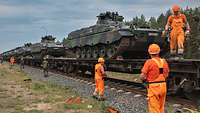 Mehrere Panzer stehen auf Bahnwaggons, davor Bahnarbeiter in orangefarbener Kleidung und Soldaten.