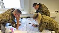 Drei Soldaten in Flecktarn-Uniform beugen sich in einem Raum über eine Landkarte, die auf einem Tisch liegt.