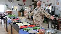 Ein Soldat steht mit ausgebreiteten Armen in einem großen Raum hinter einem Tisch, auf dem verschiedene Speisen stehen