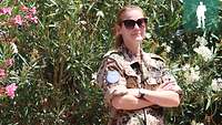 Eine Soldatin in Uniform und mit Sonnenbrille steht vor grünen Büschen. Sie hat die Arme verschränkt und lächelt.