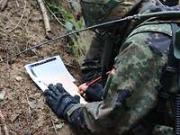 Ein Soldat mit Helm und Funkgerät auf dem Rücken schreibt auf einen beigen Zettel.