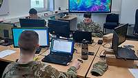 Drei Soldaten arbeiten an ihren Laptops. Im Hintergrund ist eine Karte mit farbigen Wetterdaten.