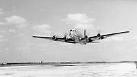 Schwarzweißaufnahme: Ein Transportflugzeug C-54 Skymaster fliegt wenige Meter über einer Startbahn 