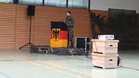 Ein Soldat steht an einem Rednerpult mit Deutschlandfahne und spricht in ein Mikrofon. 