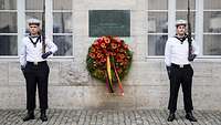 Zwei Soldaten stehen neben einer Gedenktafel und einem Blumenkranz, angebracht an einer Gebäudewand