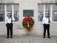 Zwei Soldaten stehen neben einer Gedenktafel und einem Blumenkranz, angebracht an einer Gebäudewand