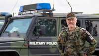 Ein Mann in Uniform steht vor einem Militärfahrzeug mit Blaulicht und lächelt in die Kamera.