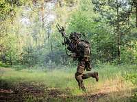 Ein Soldat rennt mit seinem Gewehr im Hochanschlag über eine Freifläche im Wald. 