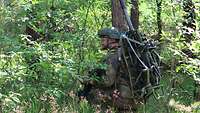 Ein Soldat kniet im Wald, auf seinem Rücken transportiert er eine sehr große und schwere Waffe.