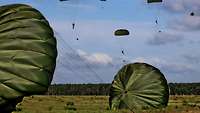 Zwei Soldaten liegen mit ihren Fallschirmen am Boden, in der Luft sind noch weitere Fallschirme.