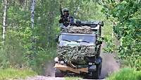 Ein Klein-Lkw fährt im Wald. Ein Soldat ragt über das Dach hinaus und beobachtet die Umgebung.